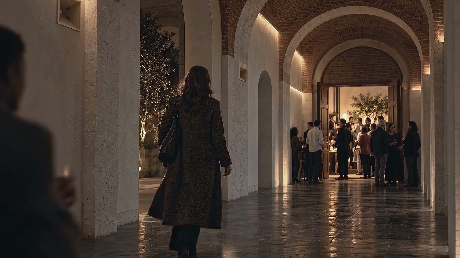 A guest walking down a long stone corridor at night towards a crowded, glowing room